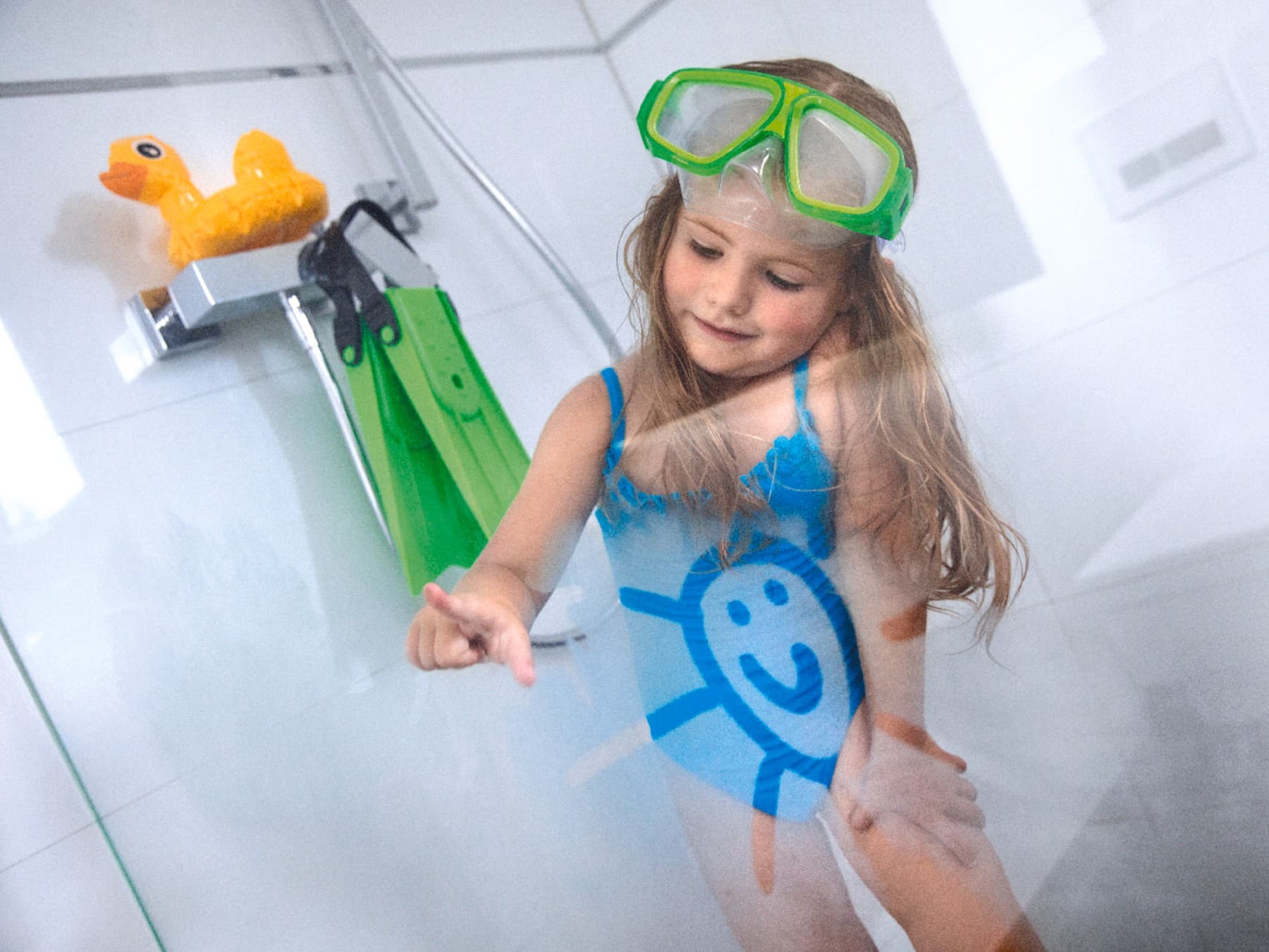 Ein Mädchen im blauen Badeanzug mit Taucherbrille und Flossen in einer Dusche mit beschlagener Duschwand. Auf die Duschwand malt sie eine lachende Sonne.