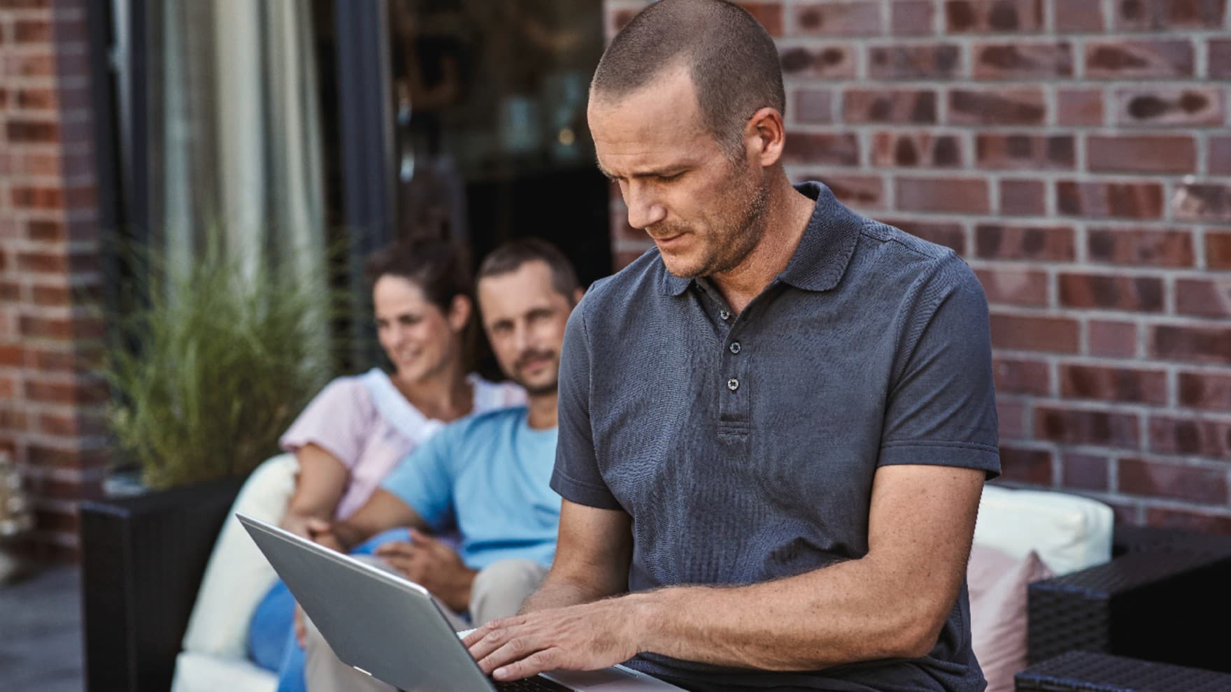 Ein Fachhandwerker sitzt draußen am Laptop und nimmt die letzten Einstellungen an der Klimaanlage vor. Die Hausbesitzer sitzen im Hintergrund.