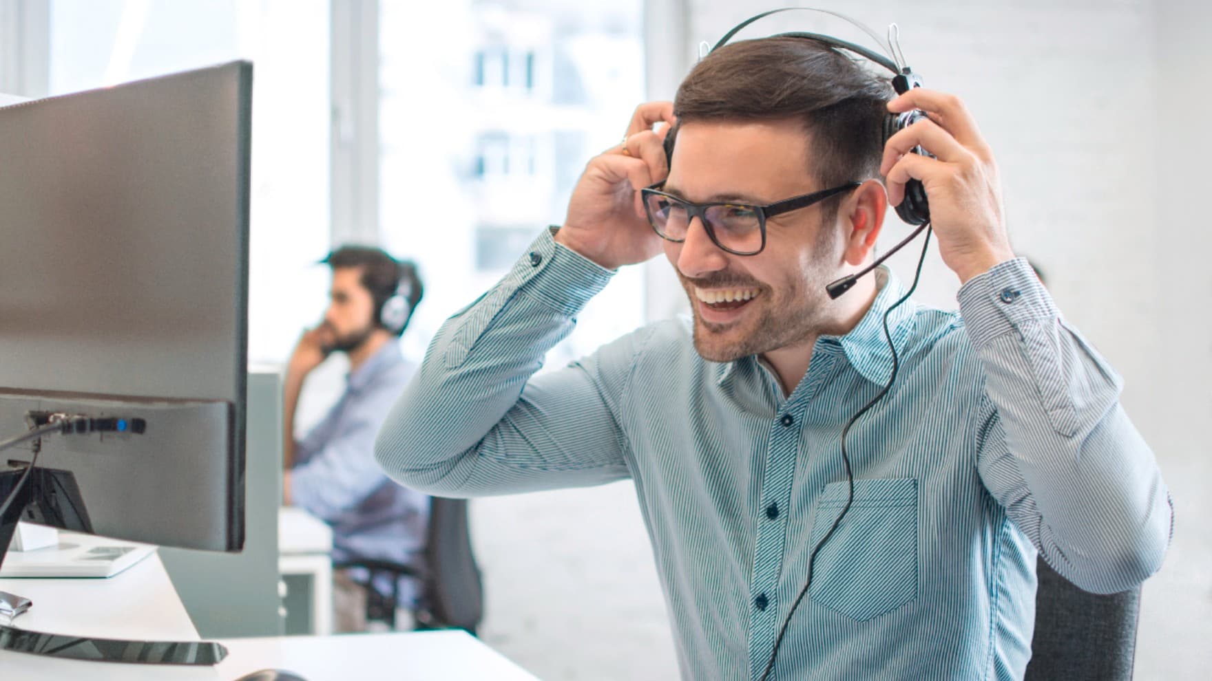 Ein Mann sitzt vor einem Computer und setzt sich lächelnd ein Headset auf.