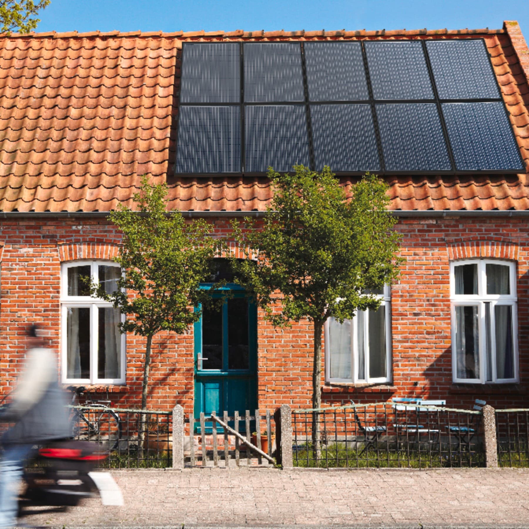 Ein altes Backsteingebäude von vorne im Sonnenlicht mit Vorgarten, auf dessen Dach sich eine Photovoltaik-Anlage befindet.