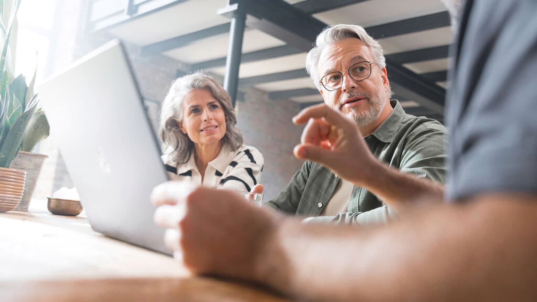 Eine Frau und ein Mann sitzen in einer Beratungssituation an einem Tisch, vor dem Mann steht ein aufgeklappter Laptop
