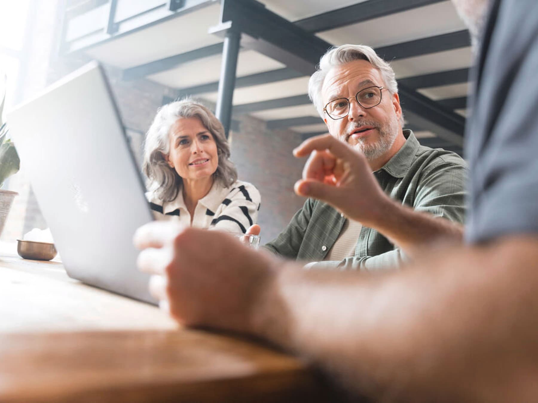 Ein Mann und eine Frau mit grauen Haaren sitzen mit einer dritten, fast nicht sichtbaren, Person an einem Tisch und haben den Blick ihm zugewandt.