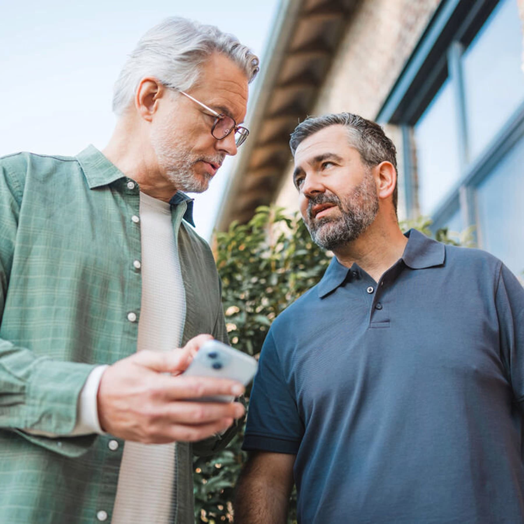 Zwei Männer von vorne unten fotografiert, stehen in ein Gespräch vertieft vor einem Haus. Der linke Mann hält ein Smartphone in der Hand, beide Männer schauen sich an.