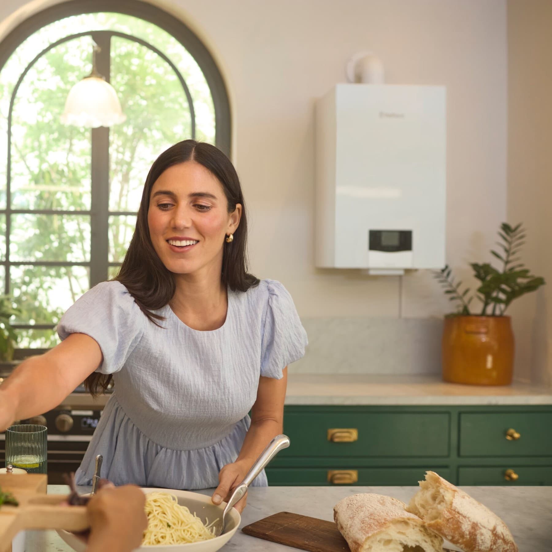 Eine Frau steht in einer gemütlich eingerichteten Küche und richtet Essen an. Hinter ihr sieht man eine zweite Küchenzeile, über der neben einem Rundfenster eine Vaillant Gasheizung hängt.