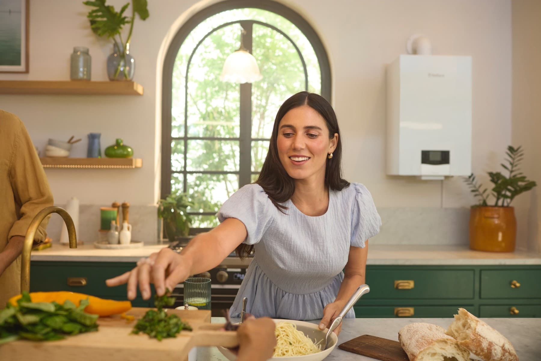Frau steht in der Küche an einer Kücheninsel und richtet Essen an. Im Hintergrund ist eine zweite Küchenzeile mit Schränken und Spüle zu sehen. Eine Vaillant Gasheizung hängt rechts im Bild über der Küchenzeile.