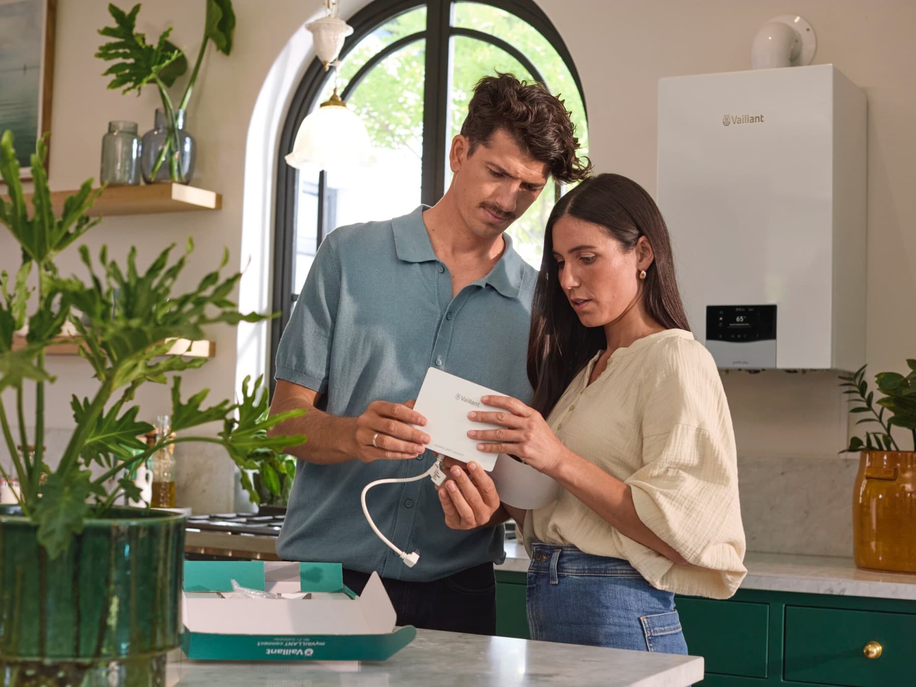 Ein Mann und eine Frau stehen gemeinsam in einer Küche und schauen sich ein technisches Gerät an. Hinter ihnen hängt eine Vaillant Brennwertheizung an der Wand neben einem Fenster.