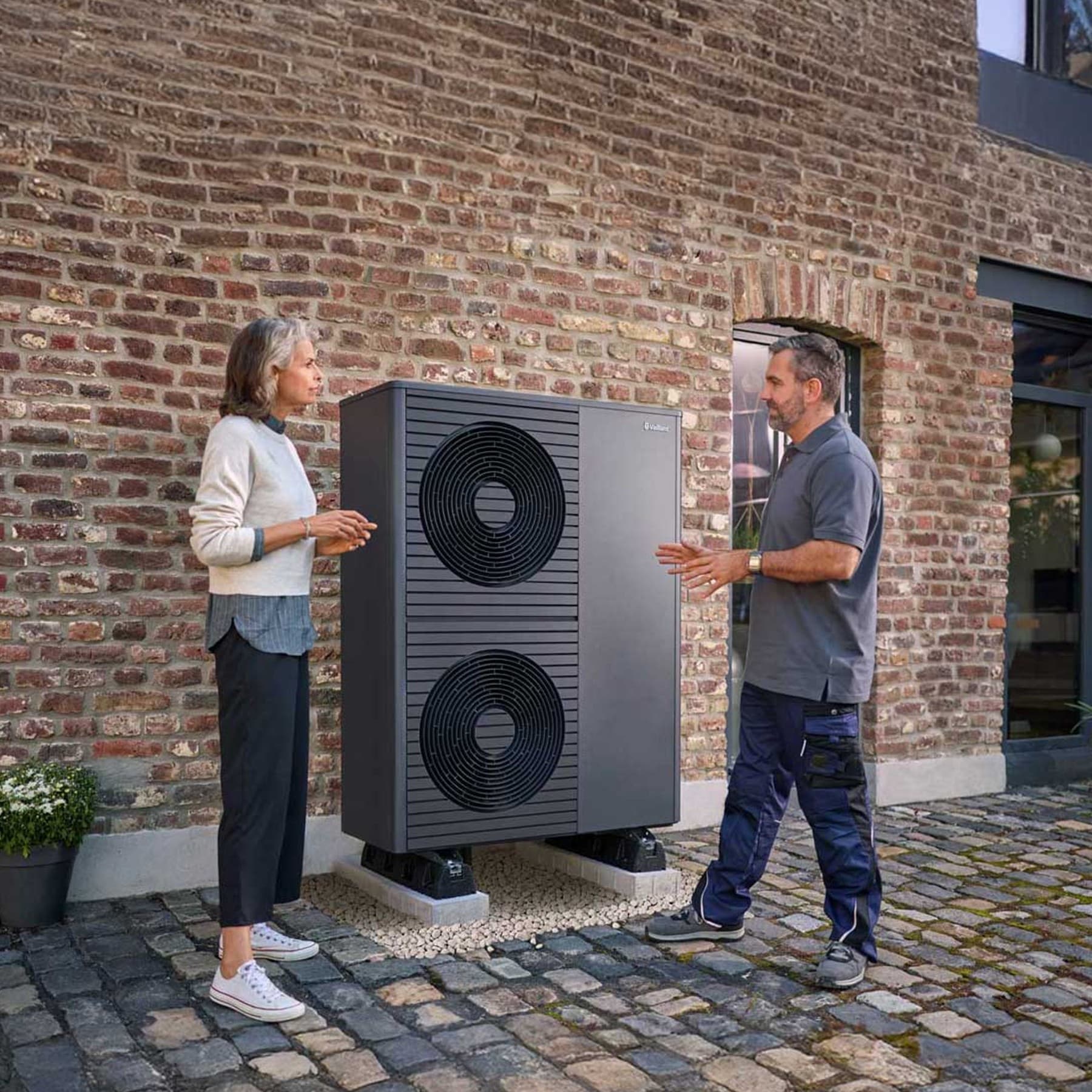 Frau steht mit Handwerker vor einem Backsteinhaus neben einer doppelstöckigen Vaillant Wärmepumpe aroTHERM plus
