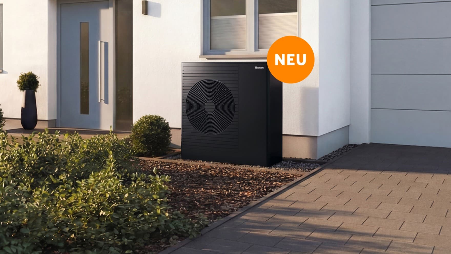 Die Vaillant aroTHERM pro steht vor einem Haus unter dem Fenster direkt neben der Eingangstür. An der Wärmepumpe ist ein orangefarbener Störer mit dem Wort "NEU".